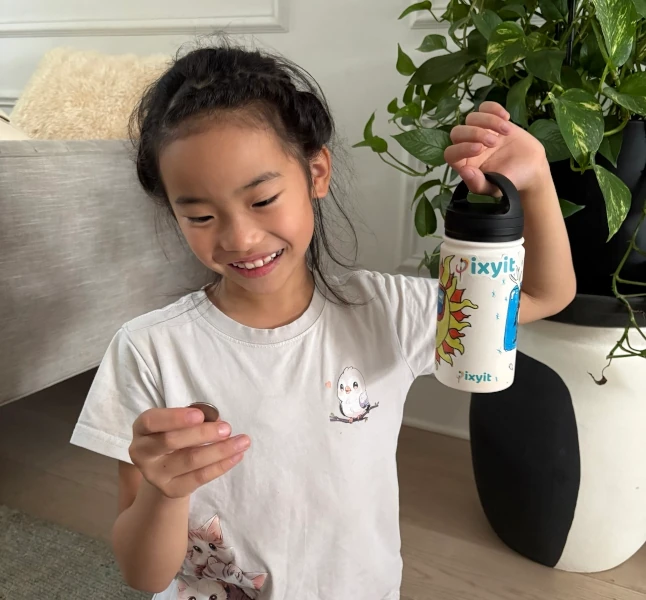 A smiling girl holding a coin and a pixyit water bottle printed with a child's sun drawing